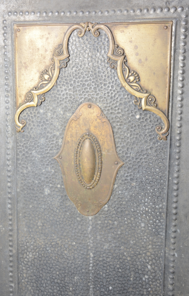 Skjermbrett av grått metall bestående av tre rektangulære paneler som hver står på to føtter. Panelene har hamret overflate og dekor i form av perlerammer. Det midterste panelet har felt med gult metall som dekorerer overflaten: de øvre hjørnene med C-former og blomster, samt en, oval dekorativ form i midten.