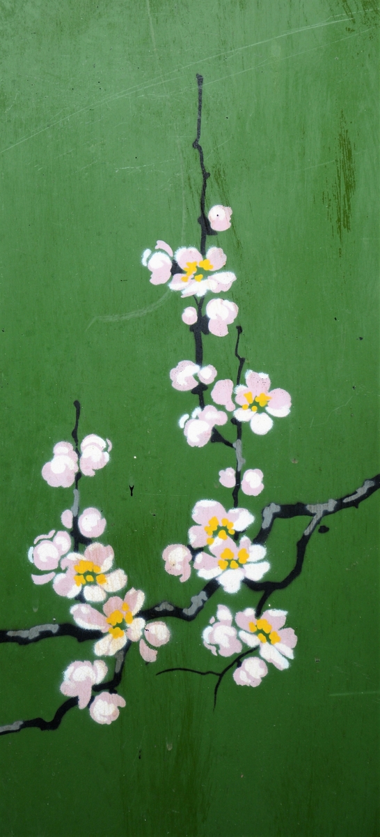 Svart skjermbrett av metall bestående av tre rektangulære paneler som hver står på to føtter. Panelene er dekorert med påmalt dekor i form av grønt vindu med grener med hvite blomster, samt sommerfugl og fugl, innrammet av tre tynne, gullborder.