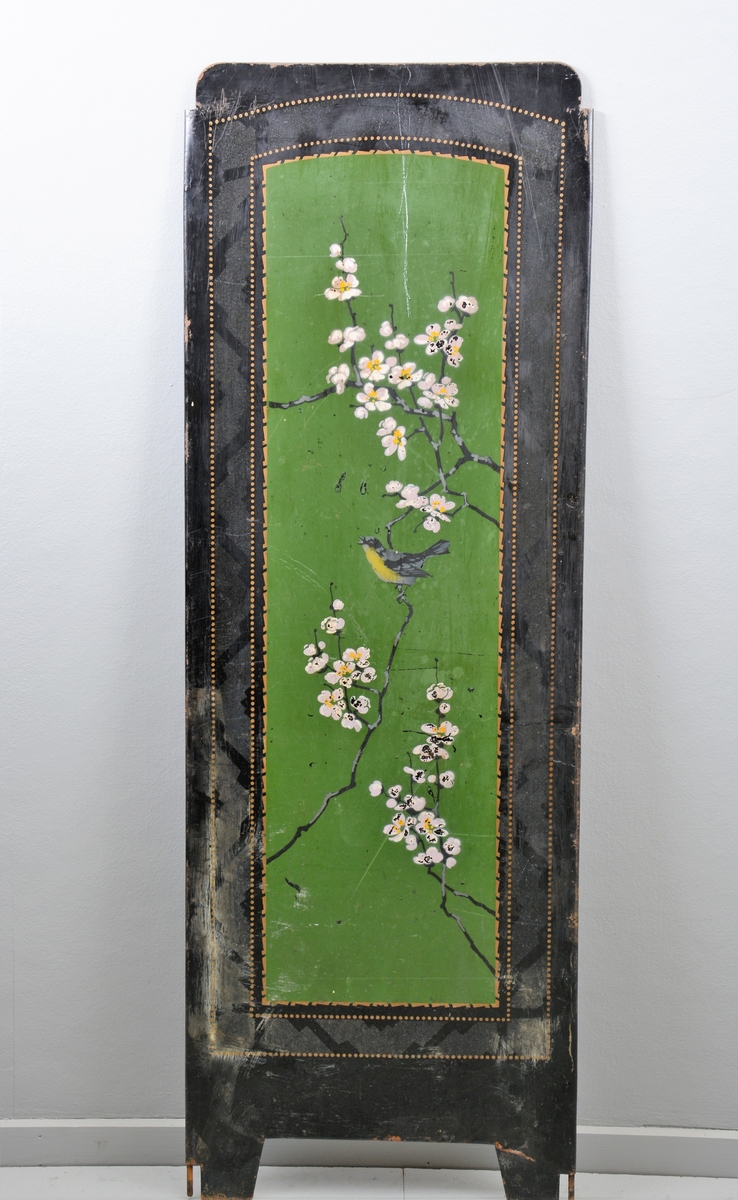 Svart skjermbrett av metall bestående av tre rektangulære paneler som hver står på to føtter. Panelene er dekorert med påmalt dekor i form av grønt vindu med grener med hvite blomster, samt sommerfugl og fugl, innrammet av tre tynne, gullborder.