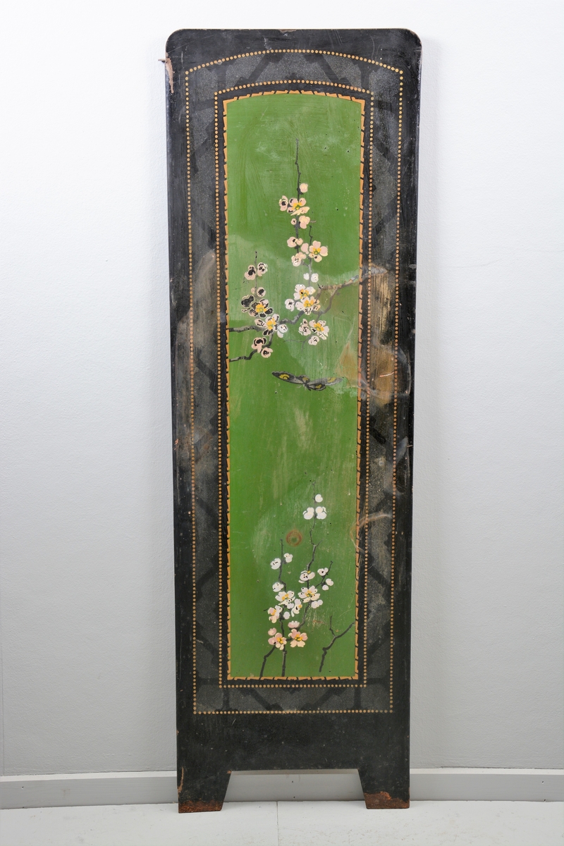 Svart skjermbrett av metall bestående av tre rektangulære paneler som hver står på to føtter. Panelene er dekorert med påmalt dekor i form av grønt vindu med grener med hvite blomster, samt sommerfugl og fugl, innrammet av tre tynne, gullborder.