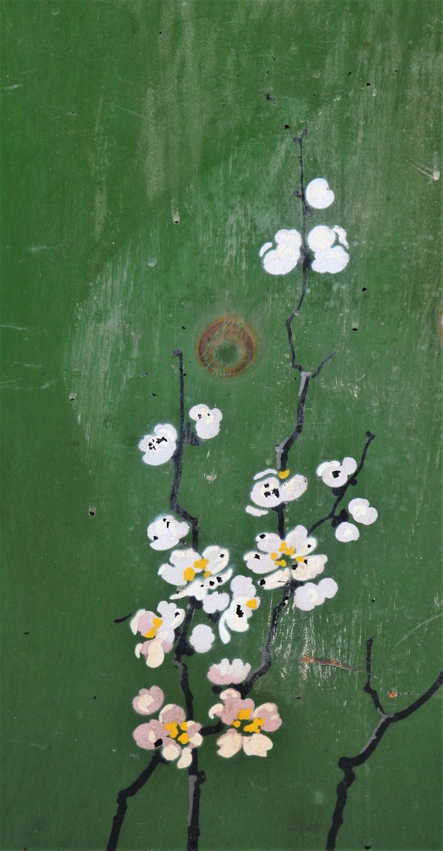 Svart skjermbrett av metall bestående av tre rektangulære paneler som hver står på to føtter. Panelene er dekorert med påmalt dekor i form av grønt vindu med grener med hvite blomster, samt sommerfugl og fugl, innrammet av tre tynne, gullborder.