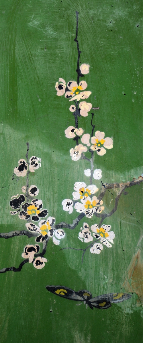 Svart skjermbrett av metall bestående av tre rektangulære paneler som hver står på to føtter. Panelene er dekorert med påmalt dekor i form av grønt vindu med grener med hvite blomster, samt sommerfugl og fugl, innrammet av tre tynne, gullborder.