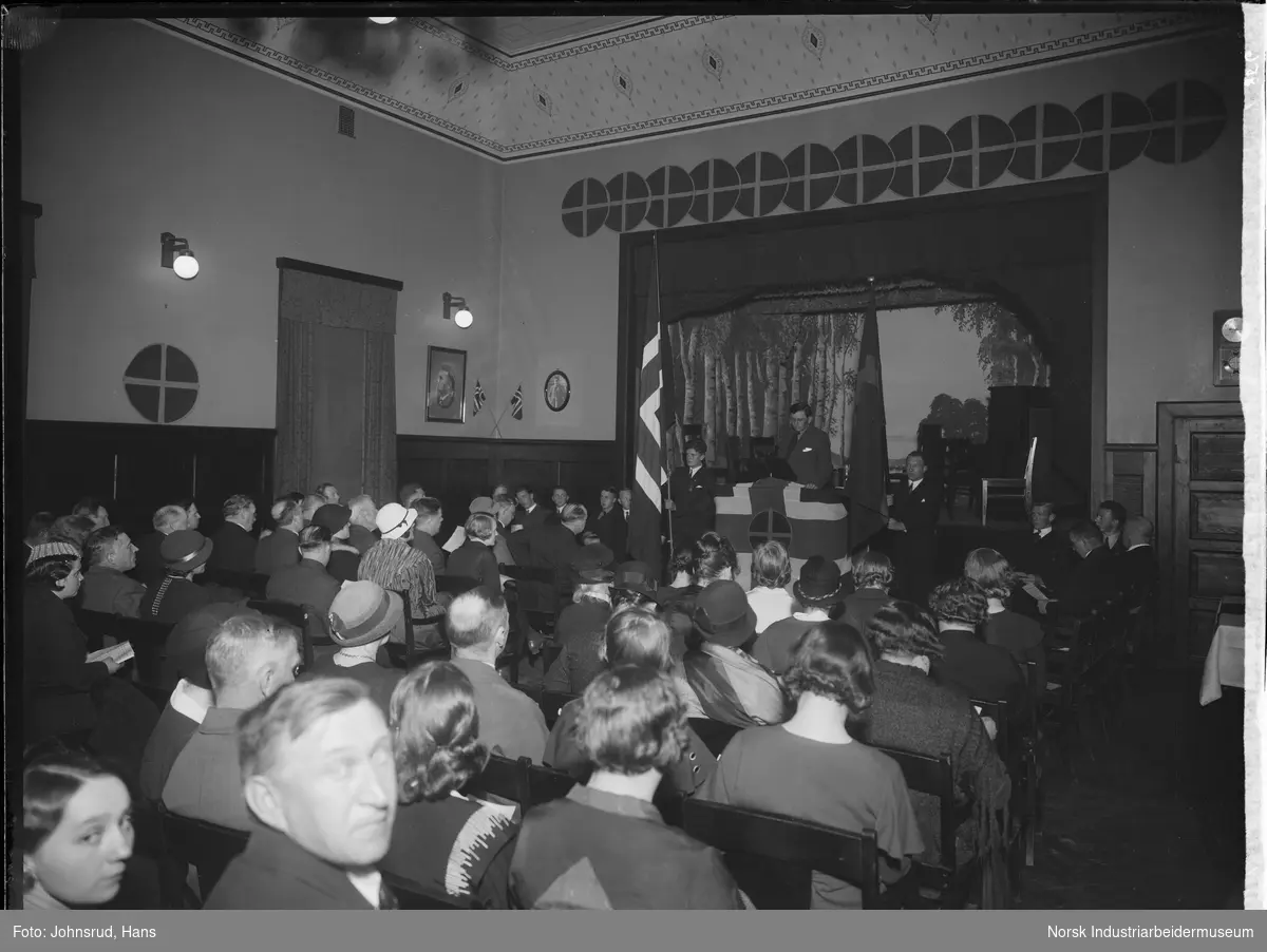 Flagginvielse ved Nasjonal Samlings møte i Godtemplarhuset i Notodden, 1934. Menn og kvinner sittende i lokale, logo for Nasjonal samling hengende rundt i lokalet. Mann holder norsk flagg ved siden av talerstolen.