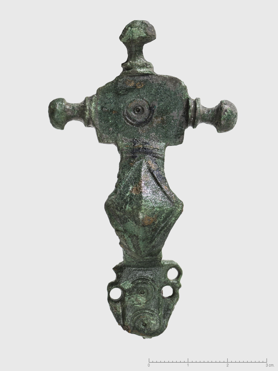 "Mindre korsformet bronsespenne, hvis øvre del med bøylen står nærmest Shetelig l. c. fig. 173, bøylen dog bredere som l. c. fig. 176. Nederste del er avbrukket ytterst, men har plate  med to profildyr, ett på hver side og to punktcirkler på den flate fotplate. Lengden av den bevarte del er 8,3 cm." Ny beskrivelse etter revisjon: Korsformet spenne av bronse med nål av jern, særform. Spennen er støpt i et stykke. Stor hodeplate med sentral konsentrisk sirkel. Knoppene er flate under, med høyt hode og fure ved foten. Bøylen er bred med vinger og har et rombisk midtfelt, avgrenset i hver ende av doble, horisontale furer. Bøyleplaten har i midtfeltet to konsentriske sirkler stående over hverandre. Langs platen er det på hver side to hull, som trolig er del av stiliserte dyr avbildet i profil. Platen er avbrutt nederst, og spennens avslutning er derfor uviss. På undersiden er nåleholder, nålefestet og hele jernnålen med spiral bevart. Det er en del korrosjon rundt nålen. Stl: 82 mm, knopper stbr: 48 mm, bøyle stbr: 20 mm.