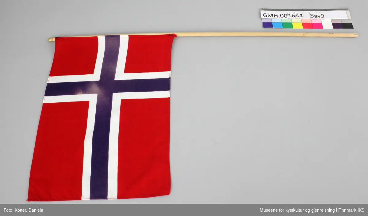 Ni norske flagg av eldre dato. Flaggene er av vevd bomullsstoff og omtrent like store. Pinnene er ulik lange, noen er knekt eller saget av, men alle har en kule, opprinnelig med gull maling på toppen.