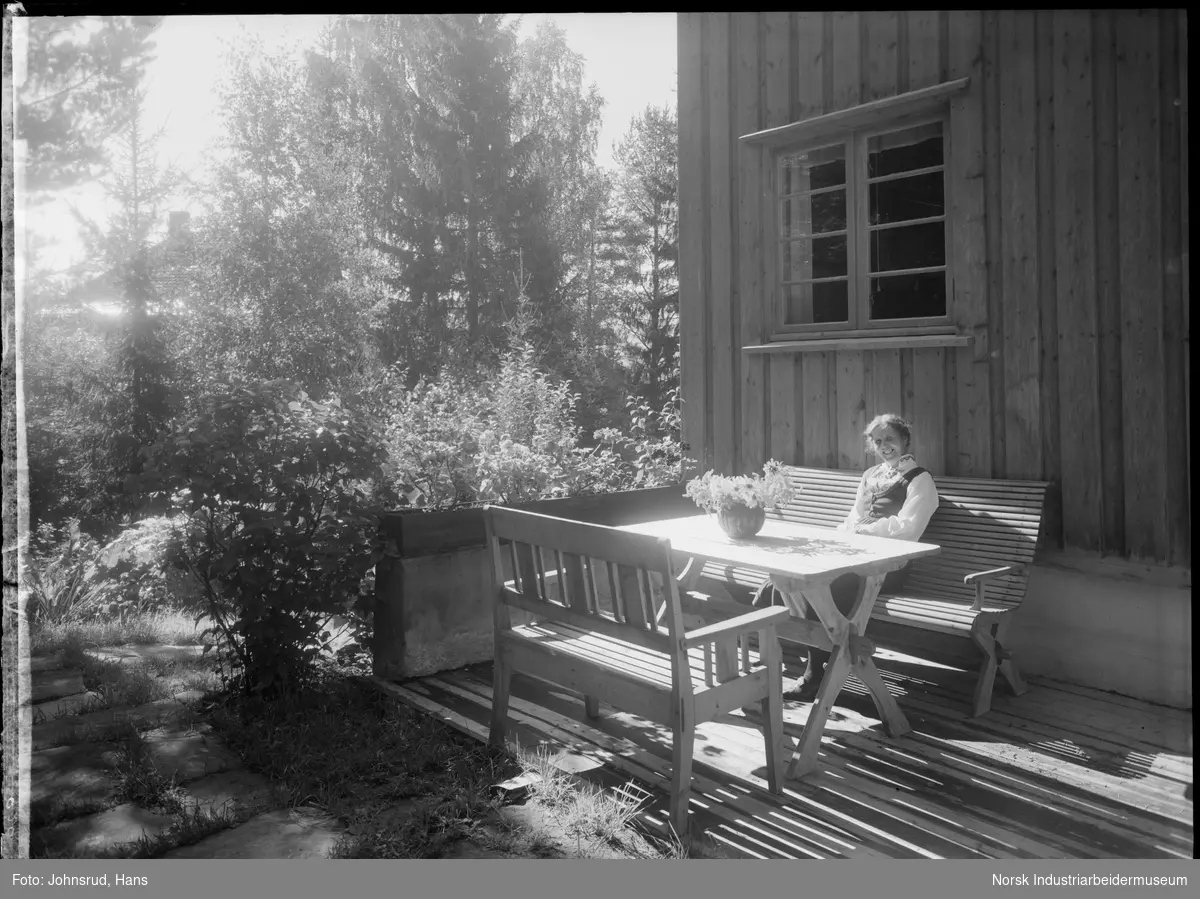 Kvinne i bunad sittende i verandamøbler foran bolighus. Blomst på bordet foran kvinnen.