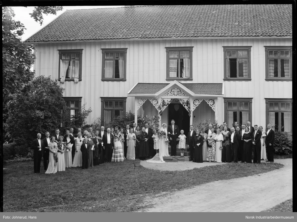 Brudepar foran bolighus med brudefølge. Brudgom er Halvor Holta. Bolighusets inngangsparti er dekorert med snekkerglede.