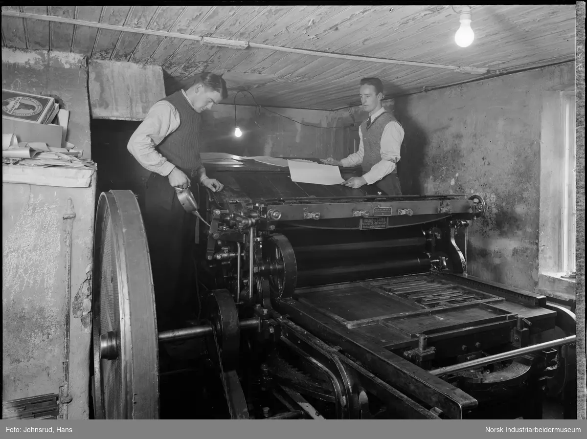 To menn arbeider på trykkpressen til Telen. En av mennene smører maskindelene, den andre står med papir på oversiden av maskinen.