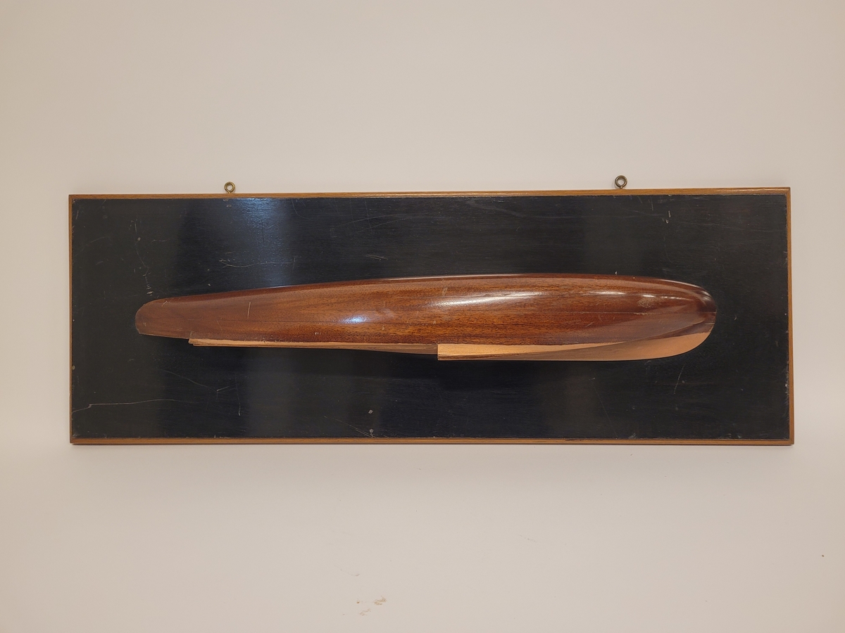 Halvmodell av motorbåt, racer. Byggd i block av trä visande styrbords sida. Skrov av mahogny med kopparfärgad botten. Monterad på svart polerad platta av mahogny med ljus ram.