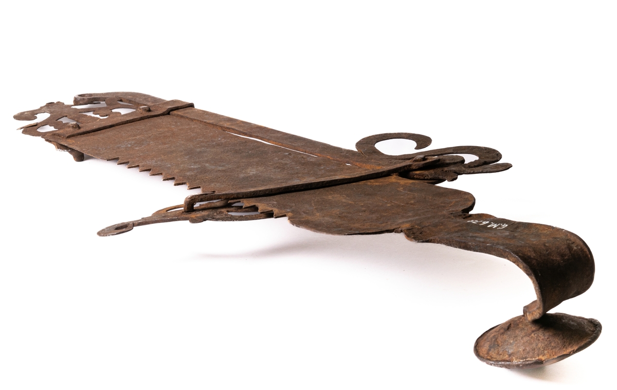 Järnföremål, okänd användning. Möjligen slangringsregel till stor ugn. Märkt 1799.
Avlångt med brutna ornament vid ändanrna liknande krona och fågelfigur.
Till vänster ett kraftigt knoppformigt handtag. Framsidan täckt av ristade ornament: slingor med glesa tvärstreck, blommor, m.m.
Otydligt smedsmärke, se skiss.
