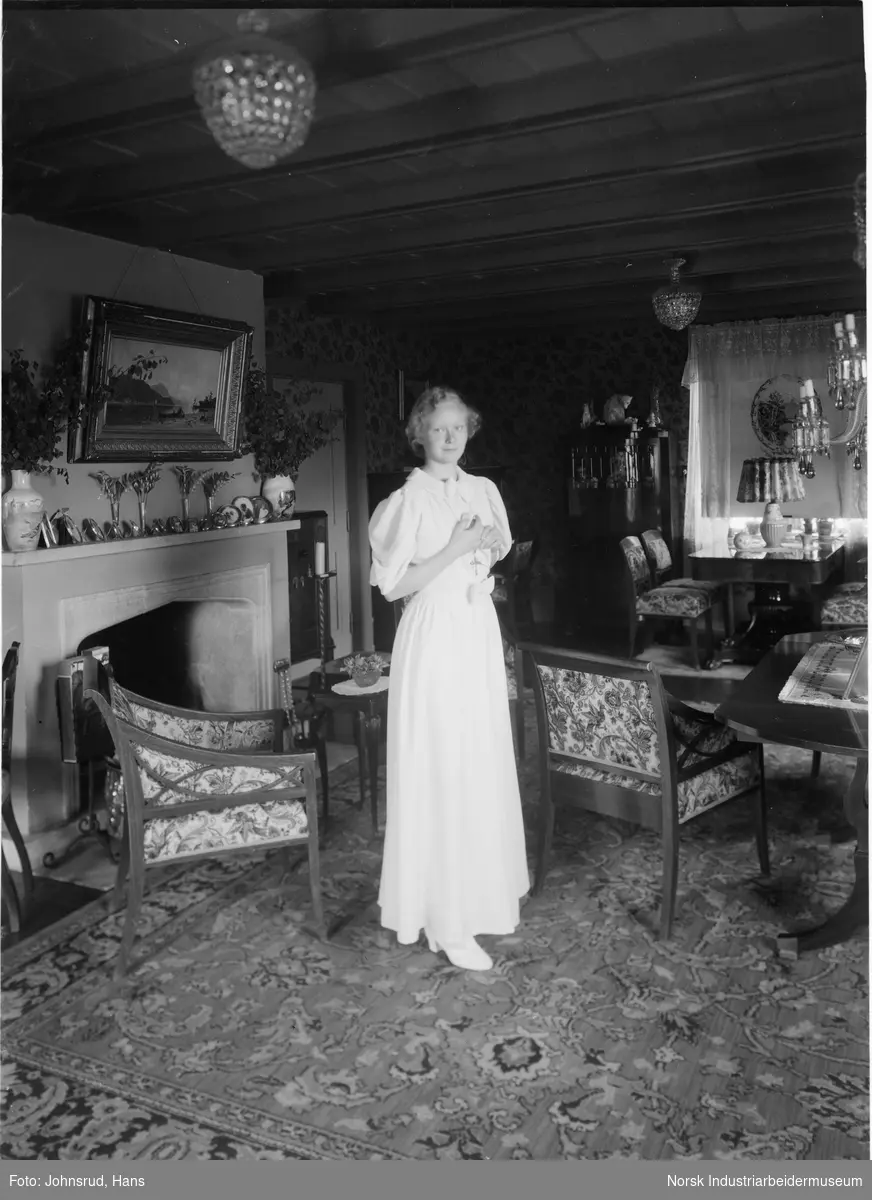 Kvinne i lys kjole oppstilt i stue. Stuen er møblert med to spisestuemøblement og peis. På peishyllen er det oppstilt flere portrettfotografier i ramme.
