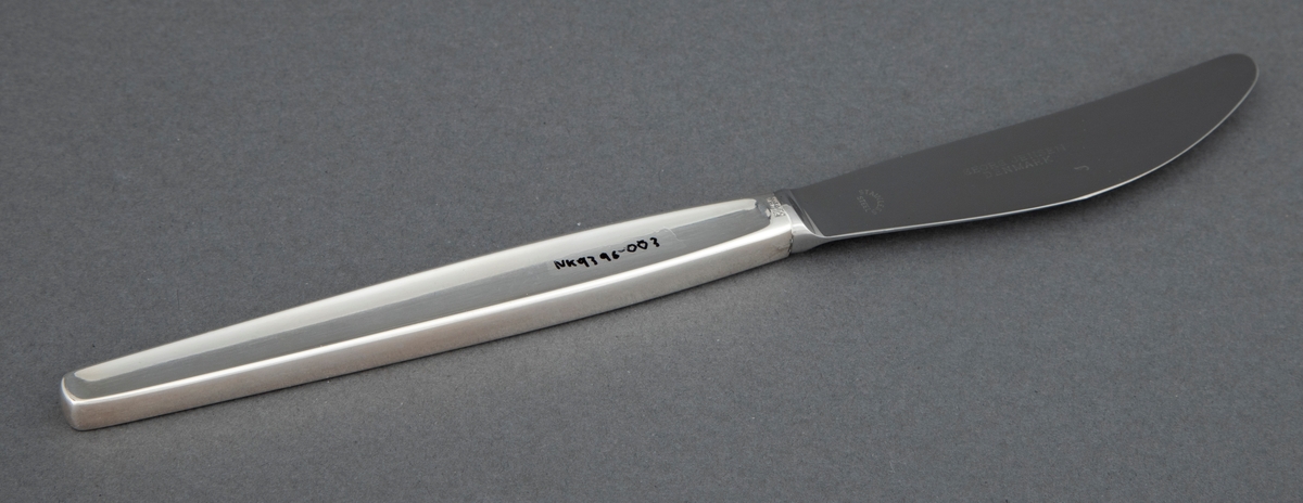 Kniv i sølv. Skaftet har rektangulært snitt, og i hele skaftlengden en ergonomisk og dekorativ forsenkning i midten på to sider. Bladet er i stål. og har buet egg.