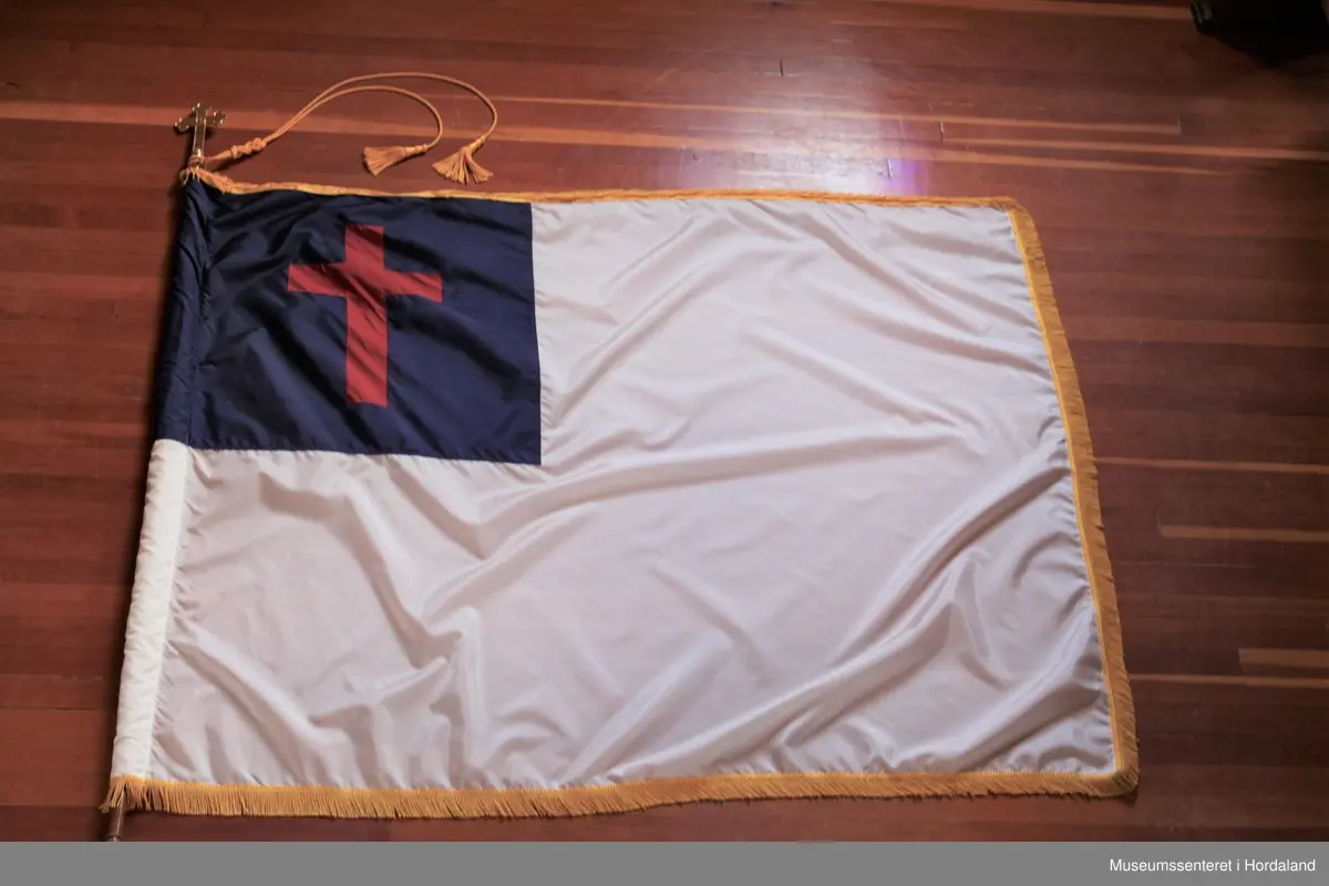 Rektangulært, hvitt flagg med et firkantet blått felt i øvre venstre hjørne med et rødt kors. Rundt flagget er en gul frynsekant. Flagget er festet til en paradestang av tre med et gyllent kors på toppen. Under dette korset er en gul snor knytt sammen ved stangen, den deler seg og ender i to dusker. Selve stangen ender i et messingledd som kan festes til en forlengelse av stangen.