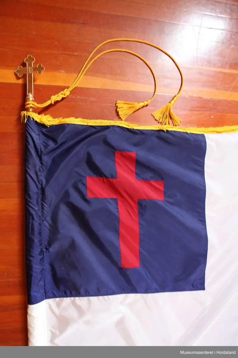 Rektangulært, hvitt flagg med et firkantet blått felt i øvre venstre hjørne med et rødt kors. Rundt flagget er en gul frynsekant. Flagget er festet til en paradestang av tre med et gyllent kors på toppen. Under dette korset er en gul snor knytt sammen ved stangen, den deler seg og ender i to dusker. Selve stangen ender i et messingledd som kan festes til en forlengelse av stangen.