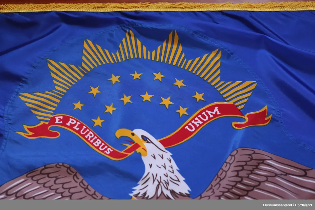Flagg på stang. Flagget er rektangulært med den amerikanske ørna, hvithodehavørn, på blå bakgrunn. Ørna er vist med utstrakte vinger, skjold på brystet, den bærer olivenkvist i den ene kloa og piler i den andre kloa, hodet er i profil, og den holder et rødt bånd i nebbet med påskriften: "E PLURIBUS UNUM" ("av mange, en"). 13 gule stjerner er fordelt på to rader i bue over hodet til ørna (de representerer de opprinnelige delstatene i USA). Over disse er en bue med 7 gule trekanter. Under ørna er et rødt bånd kantet med gul ornamentikk og påskriften: "NORTH DAKOTA" i hvitt (delstaten). Rundt flagget er en gul frynsekant. Flagget er festet til en paradestang av tre med gyllen kule og spyd på toppen.