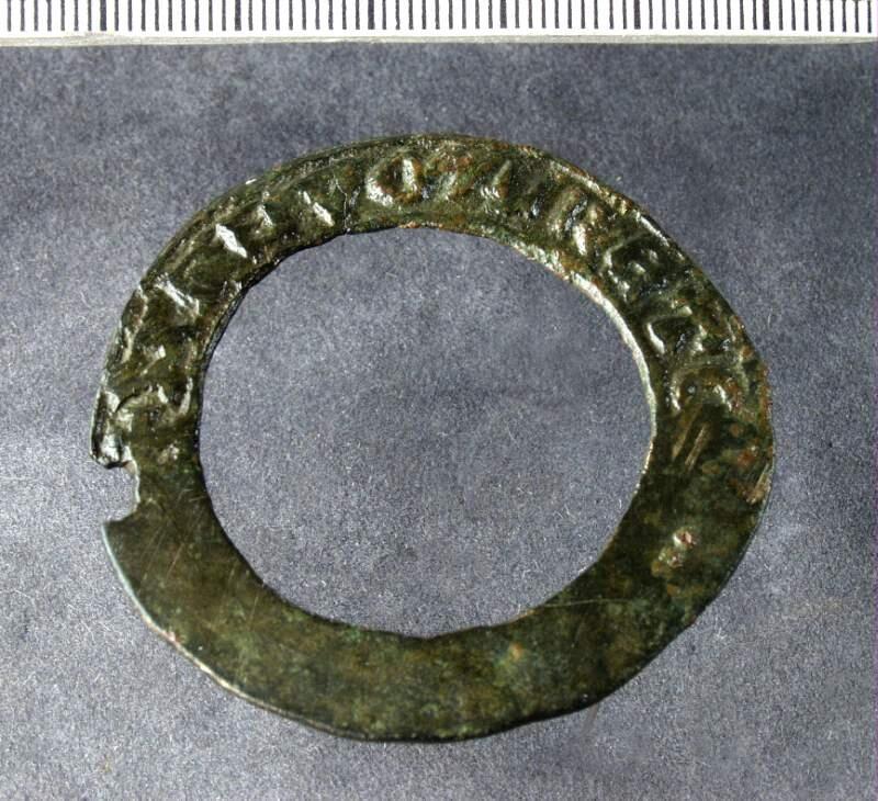 Ringspänne av platt sluten ten, ena halvan har inskrift i relief, orginalkatalogen hänvisar till a 1175:, ett "smycke" med inskrift "ave maris",( föremålet a 1175: saknas  i ark mag) texten på ovanstående spänne svårtydd, kan ev vara samma, enligt orginalkatalogen "funnen 1881 vid odling i  sigsarve-stenstugu-gärde i närheten av viges ödeslott,"
av koppar, ärgig, 2 tum i tvärmått, funnen 1881 vid odling i  sigsarve-stenstugu-gärde i närheten av viges ödeslott, med  inskrift. Se nr: 1175.