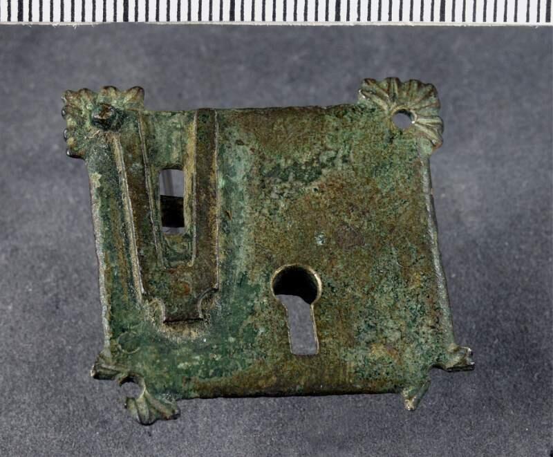 Beslag till lås av brons, närmast kvadratiskt med små bladliknande utskott i hörnen - de två nedre skadade. Kvarvarande nit för fastsättning. Litet nyckelhål, på baksidan kvarvarande låsmekanism
Av mässing, 11/4 tum i fyrkant