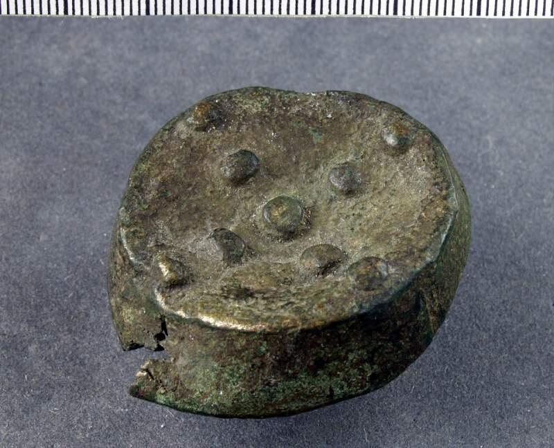 Dosformigt spänne av tidig typ, ovansidan ornerad med nio knoppar , kraftigt deformerat, nål saknas
av brons, rund.