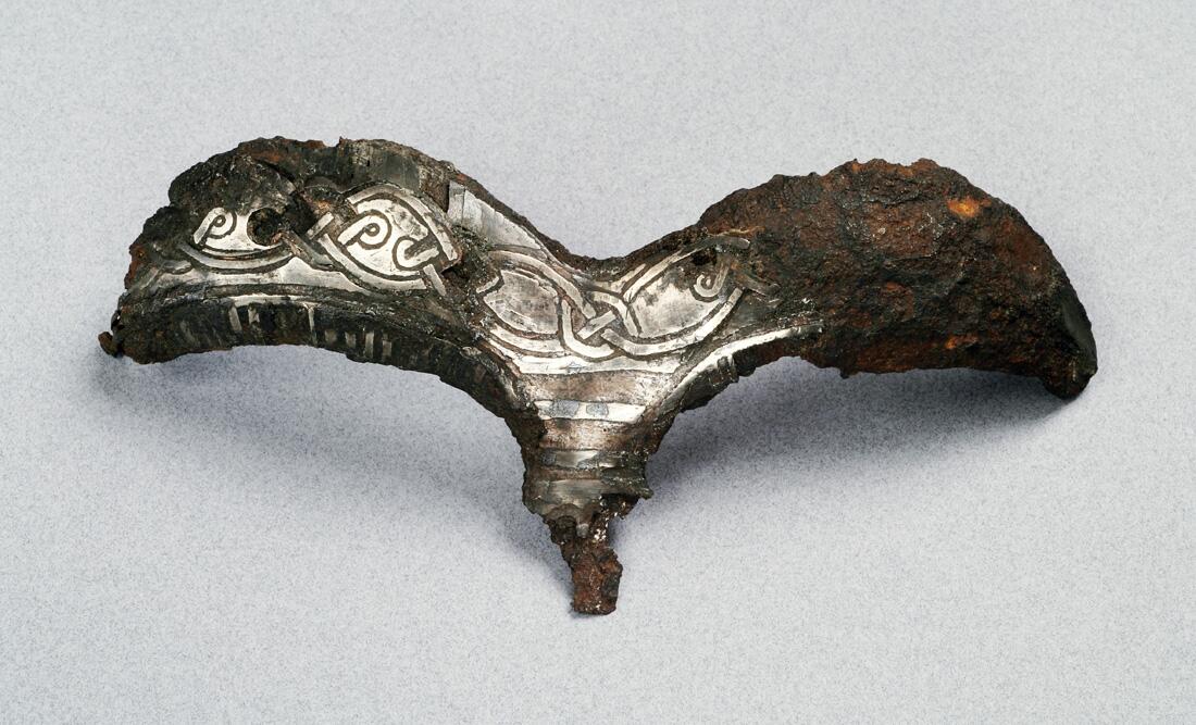 Detalj av hjälm. Av järn  närmast triskelformat skydd för ögonbryn  och näsa. Dekor av drakslingor i silver    kompl okt 1006/ml
i form av ögonbryn, av järn med drakslingor i silver. Funnen i lokrume.