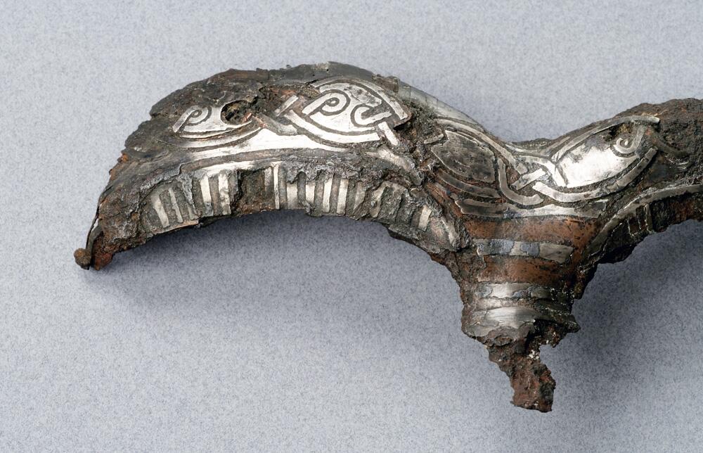 Detalj av hjälm. Av järn  närmast triskelformat skydd för ögonbryn  och näsa. Dekor av drakslingor i silver    kompl okt 1006/ml
i form av ögonbryn, av järn med drakslingor i silver. Funnen i lokrume.