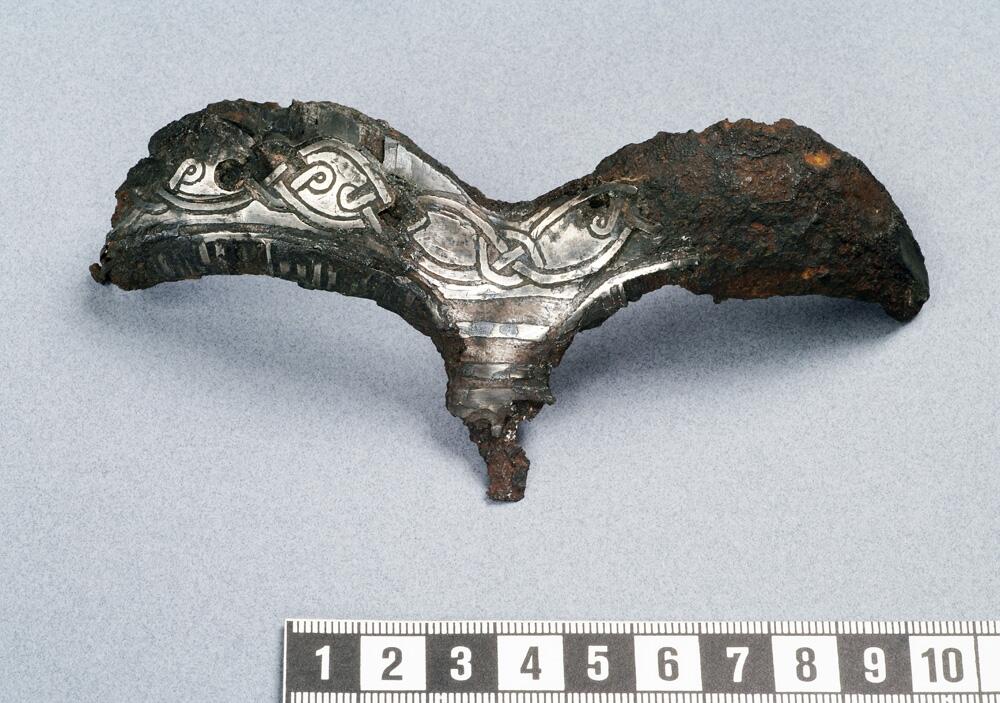 Detalj av hjälm. Av järn  närmast triskelformat skydd för ögonbryn  och näsa. Dekor av drakslingor i silver    kompl okt 1006/ml
i form av ögonbryn, av järn med drakslingor i silver. Funnen i lokrume.