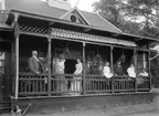 Makarna Hydén med döttrar och gäster vid sommarbostaden Talludden i Sandvik. Upplysningsvis uppfördes huset ursprungligen på den så kallade Kreatursvallen i Linköping i samband med 1898 års lantbruksmöte i staden, men flyttades därefter till en natursköna plats med utsikt över sjön Roxen. På verandan ser vi från vänster Gottfrid Söderberg, svåger respektive bror till makarna Hydén, följd av fru Maria Sofia Hydén, född Söderberg. Mot stolpen lutar sig Axelina Lundblad, dotter till den äldre mannen mellan barnen, kamrer Axel Lundblad. Barnen är makarna Hydéns döttrar Anna och Lisa. Längst till höger i bild sitter fadern, järnhandlare Mauritz Hydén. Fotoår omkring förra sekelskiftet.