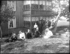 Nio kvinnor och män står och sitter i en grässlänt framför en inglasad veranda i två våningar med snickarglädje.