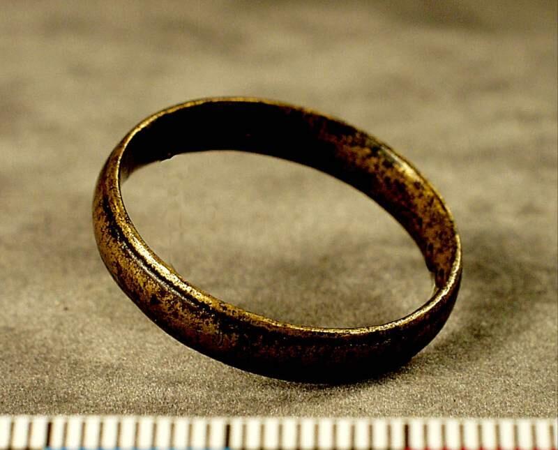 Slät något vävlvd ring ornerad med två längsgående linjer längs vardera kanten liknar mycket en modern vigselring
av brons, ornerad med två längsgående linjer, utsidan svagt välvd. Diam. 2,5 cm.