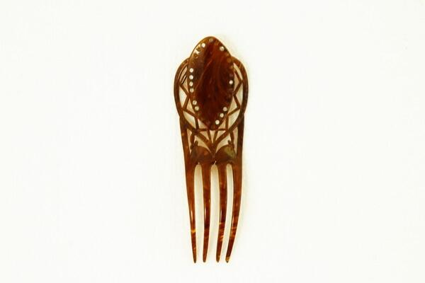 Prydnadskam av sköldpaddsimitation, (plast) med fyra tänder, hög rygg med genombruten dekor som har dekorprickar av metall. Slipade glasstenar (strass) runt den ovala mittenplattan. 17 st av 30 saknas. Bredd 5,5 cm, höjd 17 cm. Ändrad av k.h & s.h Fma hgo.
av Sköldpadd, fyra tänder, hög rygg med genombruten dekor och slipade glasstenar. L. 17 cm. Br. 5,5 cm.