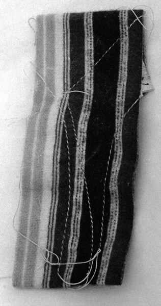 a) Symaskin av märket Husqvarna, modell V.S.4. Handdriven. Svartmålad på träplatta. Nersänkt låda med tillbehör. b) Huv av ekfaner. Handtag på toppen. c) Skyttel av stål. Rund, spetsig i ena änden, rak i den andra. Innehåller en spole med vit tråd. d) Fem spolar av stål. Rund stav med plattor i ändarna. Olika trådrester. e) Ledpressarfot (monterad på maskinen). f) Fållvikare 2 mm. g) Fållvikare 6 mm. h) Gansapparat. i) Rynkfot .j) Linjal. Längd 5,3 cm. k) Fem symaskinsnålar i olika grovlekar. Cellofanförpackning. l) Skruvnyckel. Typ lång platt nål, bakre delen försedd med runt hål. m) Skruvmejsel av stål. Fäste av svart bakelit. n) Borste av dubbelvriden ståltråd med en borste i ena änden. o) Skruvmejsel av stål. Fäste av svart bakelit. p) Provlapp av bomullsflanell. För kontroll av stygnställning. q) Provlapp av bomyllskypert. Till kontroll av stygnställning. r) Bruksanvisning. Tryckt broschyr på 30 sidor.