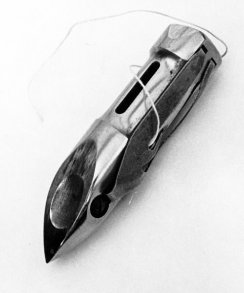 a) Symaskin av märket Husqvarna, modell V.S.4. Handdriven. Svartmålad på träplatta. Nersänkt låda med tillbehör. b) Huv av ekfaner. Handtag på toppen. c) Skyttel av stål. Rund, spetsig i ena änden, rak i den andra. Innehåller en spole med vit tråd. d) Fem spolar av stål. Rund stav med plattor i ändarna. Olika trådrester. e) Ledpressarfot (monterad på maskinen). f) Fållvikare 2 mm. g) Fållvikare 6 mm. h) Gansapparat. i) Rynkfot .j) Linjal. Längd 5,3 cm. k) Fem symaskinsnålar i olika grovlekar. Cellofanförpackning. l) Skruvnyckel. Typ lång platt nål, bakre delen försedd med runt hål. m) Skruvmejsel av stål. Fäste av svart bakelit. n) Borste av dubbelvriden ståltråd med en borste i ena änden. o) Skruvmejsel av stål. Fäste av svart bakelit. p) Provlapp av bomullsflanell. För kontroll av stygnställning. q) Provlapp av bomyllskypert. Till kontroll av stygnställning. r) Bruksanvisning. Tryckt broschyr på 30 sidor.