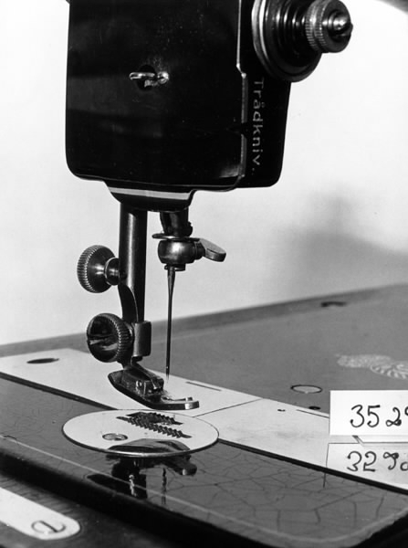 a) Symaskin av märket Husqvarna, modell V.S.4. Handdriven. Svartmålad på träplatta. Nersänkt låda med tillbehör. b) Huv av ekfaner. Handtag på toppen. c) Skyttel av stål. Rund, spetsig i ena änden, rak i den andra. Innehåller en spole med vit tråd. d) Fem spolar av stål. Rund stav med plattor i ändarna. Olika trådrester. e) Ledpressarfot (monterad på maskinen). f) Fållvikare 2 mm. g) Fållvikare 6 mm. h) Gansapparat. i) Rynkfot .j) Linjal. Längd 5,3 cm. k) Fem symaskinsnålar i olika grovlekar. Cellofanförpackning. l) Skruvnyckel. Typ lång platt nål, bakre delen försedd med runt hål. m) Skruvmejsel av stål. Fäste av svart bakelit. n) Borste av dubbelvriden ståltråd med en borste i ena änden. o) Skruvmejsel av stål. Fäste av svart bakelit. p) Provlapp av bomullsflanell. För kontroll av stygnställning. q) Provlapp av bomyllskypert. Till kontroll av stygnställning. r) Bruksanvisning. Tryckt broschyr på 30 sidor.