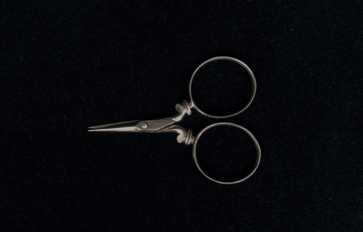 a) Syskrin av päronträ. Rektangulärt med ett metallhandtag på lockets översida och en pärlemorplatta infälld i träet under handtaget. Skrinet har fyra metallkulor som fötter. En spegel finns i lockets insida. b-c) Nåletui med lock i pärlemor med gulmetall i ett band runt etuiets lock och en stiliserad blomma, blå och grön med gul botten på etuiets framsida. d) Fingerborg av pärlemorliknande material med två gula metallringar. Höjd 2,5 cm, diameter 1,6 cm. e) Parfymflaska av slipat glas med en metallpropp. Oval tillplattad modell. Längd 5,5 cm, bredd 2,7 cm, tjocklek 0,4 cm. f) Syl av järn med pärlemorskaft. Mellan järnet och skaftet en gul metall utsmyckning. På skaftet en stiliserad blomma i blått med gul bakgrund. g) Sax av järn. Längd 5 cm, bredd 3,6 cm, tjocklek 0,2 cm. h) Sax av järn med pärlemorskaft med detaljer av gul metall. Längd 9,5 cm, bredd 3,7 cm, tjocklek 0,3 cm. i) Stoppnål av järn med dubbla ögon. Mått: längd 7 cm,  diameter 0,1 cm. j) Trådhållare av pärlemor med fyra utfällda "armar" och dekorationer i form av cirklar med mera. Längd ihopfälld 6 cm, bredd 1 cm, tjocklek 0,3 cm. 
k) Trådhållare i form av en stjärna av pärlemor med små hål i form av stjärnor. Diameter 3 cm, tjocklek 0,1 cm. l) Trådrulle i form av en stjärna av pärlemor med små hål i form av stjärnor. Mått: diameter 3 cm, tjocklek 0,1 cm. m) Trådrulle av pärlemor. Mått: längd 2,9 cm, diameter 1,5 cm. o) Insats till skrinet, trä, textil. Längd 17 cm, bredd 11,5 cm, tjocklek 0,9 cm. p) Skydd av textil som ligger mellan locket och insatsen med syutensilierna.