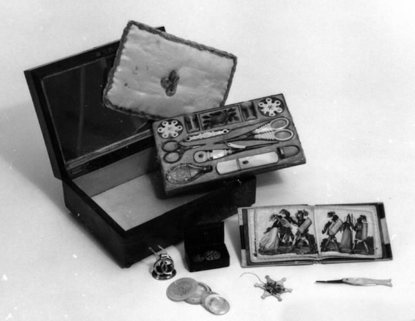 a) Syskrin av päronträ. Rektangulärt med ett metallhandtag på lockets översida och en pärlemorplatta infälld i träet under handtaget. Skrinet har fyra metallkulor som fötter. En spegel finns i lockets insida. b-c) Nåletui med lock i pärlemor med gulmetall i ett band runt etuiets lock och en stiliserad blomma, blå och grön med gul botten på etuiets framsida. d) Fingerborg av pärlemorliknande material med två gula metallringar. Höjd 2,5 cm, diameter 1,6 cm. e) Parfymflaska av slipat glas med en metallpropp. Oval tillplattad modell. Längd 5,5 cm, bredd 2,7 cm, tjocklek 0,4 cm. f) Syl av järn med pärlemorskaft. Mellan järnet och skaftet en gul metall utsmyckning. På skaftet en stiliserad blomma i blått med gul bakgrund. g) Sax av järn. Längd 5 cm, bredd 3,6 cm, tjocklek 0,2 cm. h) Sax av järn med pärlemorskaft med detaljer av gul metall. Längd 9,5 cm, bredd 3,7 cm, tjocklek 0,3 cm. i) Stoppnål av järn med dubbla ögon. Mått: längd 7 cm,  diameter 0,1 cm. j) Trådhållare av pärlemor med fyra utfällda "armar" och dekorationer i form av cirklar med mera. Längd ihopfälld 6 cm, bredd 1 cm, tjocklek 0,3 cm. 
k) Trådhållare i form av en stjärna av pärlemor med små hål i form av stjärnor. Diameter 3 cm, tjocklek 0,1 cm. l) Trådrulle i form av en stjärna av pärlemor med små hål i form av stjärnor. Mått: diameter 3 cm, tjocklek 0,1 cm. m) Trådrulle av pärlemor. Mått: längd 2,9 cm, diameter 1,5 cm. o) Insats till skrinet, trä, textil. Längd 17 cm, bredd 11,5 cm, tjocklek 0,9 cm. p) Skydd av textil som ligger mellan locket och insatsen med syutensilierna.