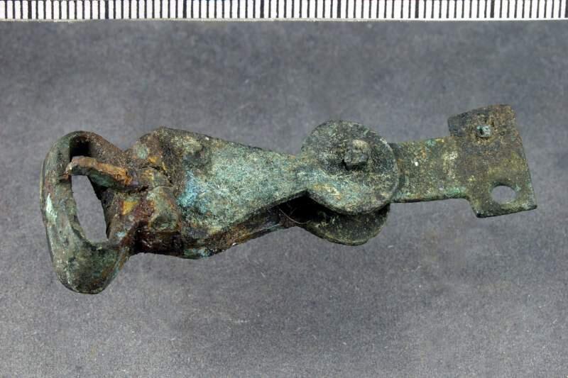 AV BRONS, MED TORNE AV JÄRN, PROFILERAD. FÄSTEBESLAGET I ETT STYCKE I SÖLJAN. BRONSBESLAG FÄSTAT MED EN BRONSNIT VID DEN CIRKLERUNDA AVSLUTNINGEN PÅ FÄSTEBESLAGET. L. 6 CM. ST. BR. 2,3 CM. FUNNEN NUMRERAD VID INV. 1968.