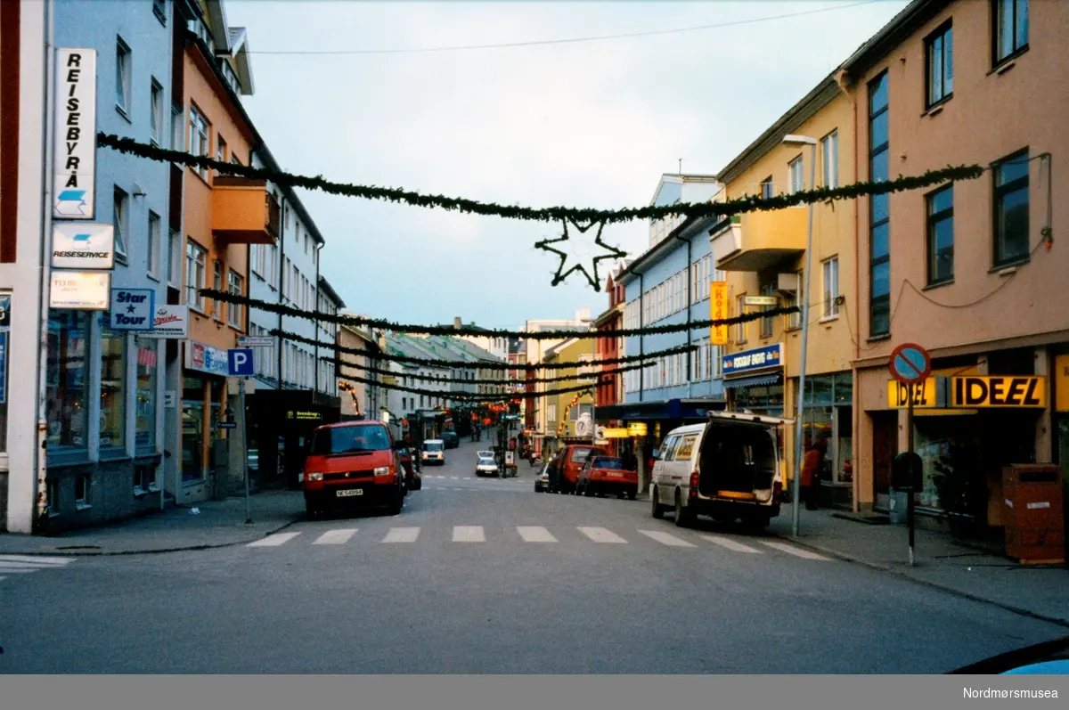 Foto fra Nedre Enggate på Kirklandet i Kristiansund. Her ser vi forretninger som for eks. Reiseservice, Ideel, Fotograf Engvig, Einar Engvig AS og Sverdrup.  Arkivskaper og giver var Stein Magne Bach ved Nordmøre museum.