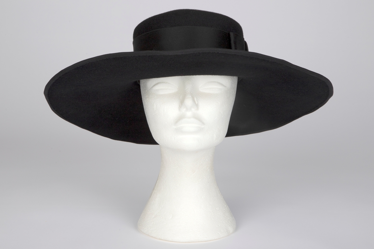 Bredbremmet filthatt med nokså flat, lav pull, formpresset av ett stykke. Nesten rettvinklet overgang mellom pull og brem. Bremmen er lett oppoverbøyd i sidene og foran, og lett skrånende midt bak. I ytterkant påsydd et smalt ripsbånd omkring en ståltråd. Pullens sider er nesten dekket av et bredt, sateng hattebånd, med dekorativ, flat sløyfe på venstre side. Midt bak er det stukket en kort hattenål, med dråpeformet endestykke av svart filt. Innvendig svettebånd av ripsbånd.