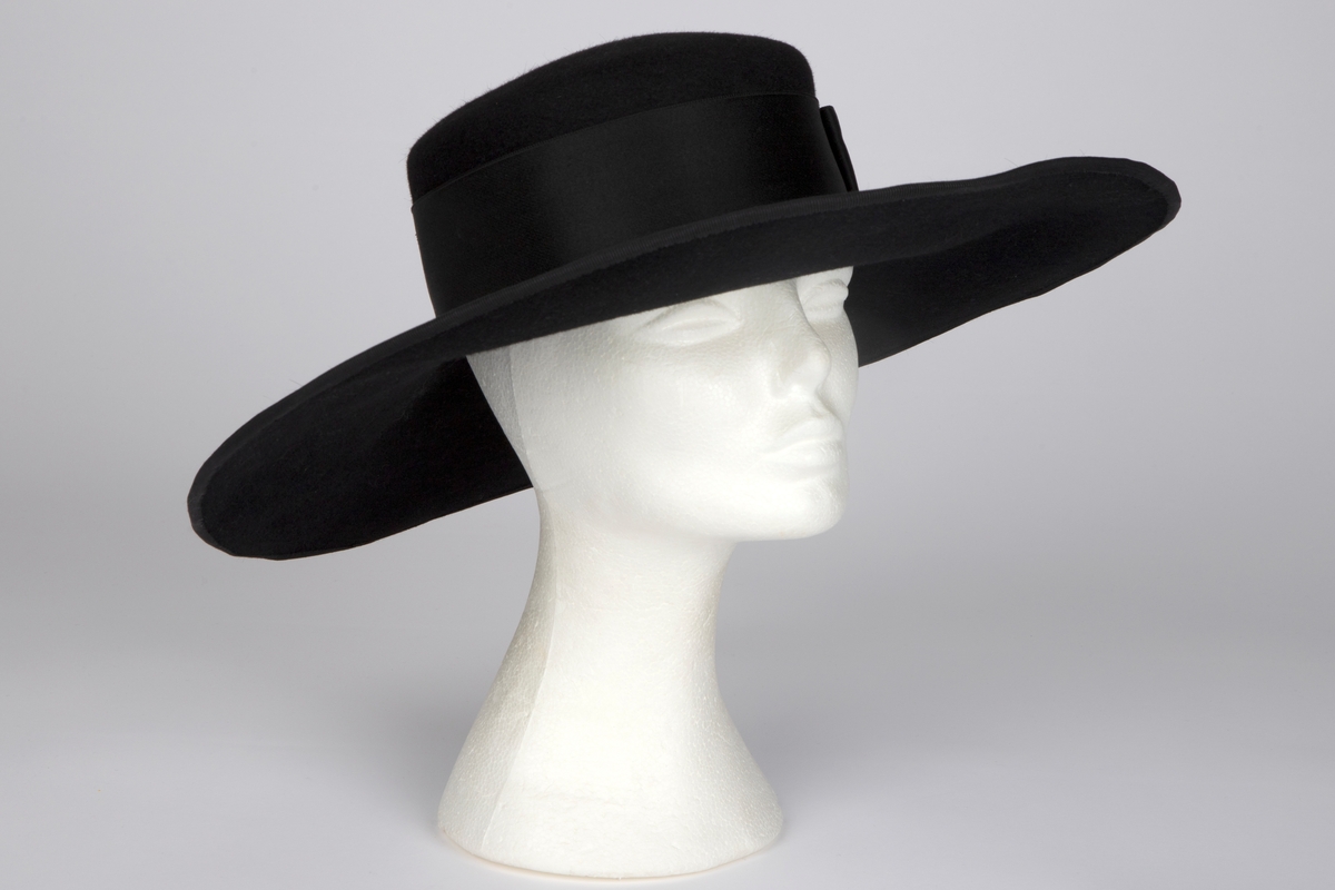 Bredbremmet filthatt med nokså flat, lav pull, formpresset av ett stykke. Nesten rettvinklet overgang mellom pull og brem. Bremmen er lett oppoverbøyd i sidene og foran, og lett skrånende midt bak. I ytterkant påsydd et smalt ripsbånd omkring en ståltråd. Pullens sider er nesten dekket av et bredt, sateng hattebånd, med dekorativ, flat sløyfe på venstre side. Midt bak er det stukket en kort hattenål, med dråpeformet endestykke av svart filt. Innvendig svettebånd av ripsbånd.