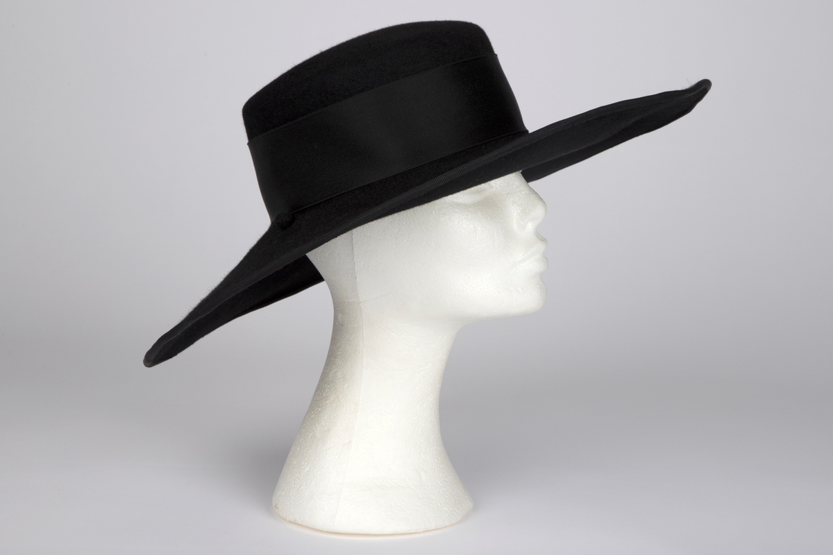 Bredbremmet filthatt med nokså flat, lav pull, formpresset av ett stykke. Nesten rettvinklet overgang mellom pull og brem. Bremmen er lett oppoverbøyd i sidene og foran, og lett skrånende midt bak. I ytterkant påsydd et smalt ripsbånd omkring en ståltråd. Pullens sider er nesten dekket av et bredt, sateng hattebånd, med dekorativ, flat sløyfe på venstre side. Midt bak er det stukket en kort hattenål, med dråpeformet endestykke av svart filt. Innvendig svettebånd av ripsbånd.