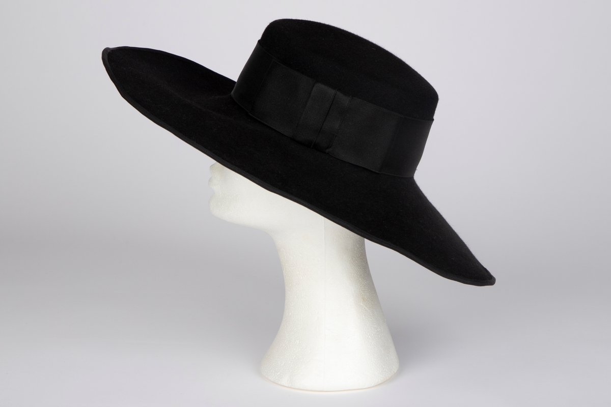 Bredbremmet filthatt med nokså flat, lav pull, formpresset av ett stykke. Nesten rettvinklet overgang mellom pull og brem. Bremmen er lett oppoverbøyd i sidene og foran, og lett skrånende midt bak. I ytterkant påsydd et smalt ripsbånd omkring en ståltråd. Pullens sider er nesten dekket av et bredt, sateng hattebånd, med dekorativ, flat sløyfe på venstre side. Midt bak er det stukket en kort hattenål, med dråpeformet endestykke av svart filt. Innvendig svettebånd av ripsbånd.