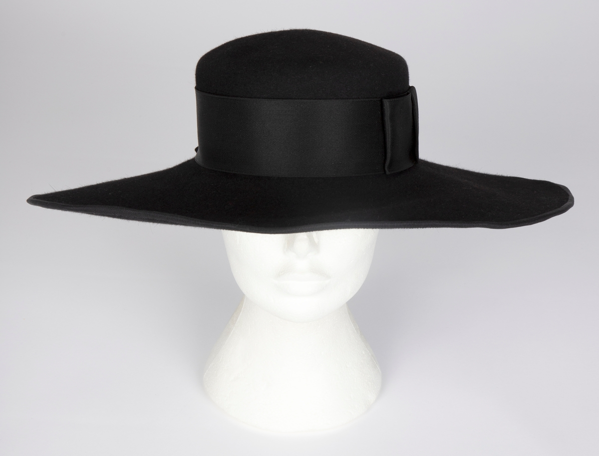 Bredbremmet filthatt med nokså flat, lav pull, formpresset av ett stykke. Nesten rettvinklet overgang mellom pull og brem. Bremmen er lett oppoverbøyd i sidene og foran, og lett skrånende midt bak. I ytterkant påsydd et smalt ripsbånd omkring en ståltråd. Pullens sider er nesten dekket av et bredt, sateng hattebånd, med dekorativ, flat sløyfe på venstre side. Midt bak er det stukket en kort hattenål, med dråpeformet endestykke av svart filt. Innvendig svettebånd av ripsbånd.