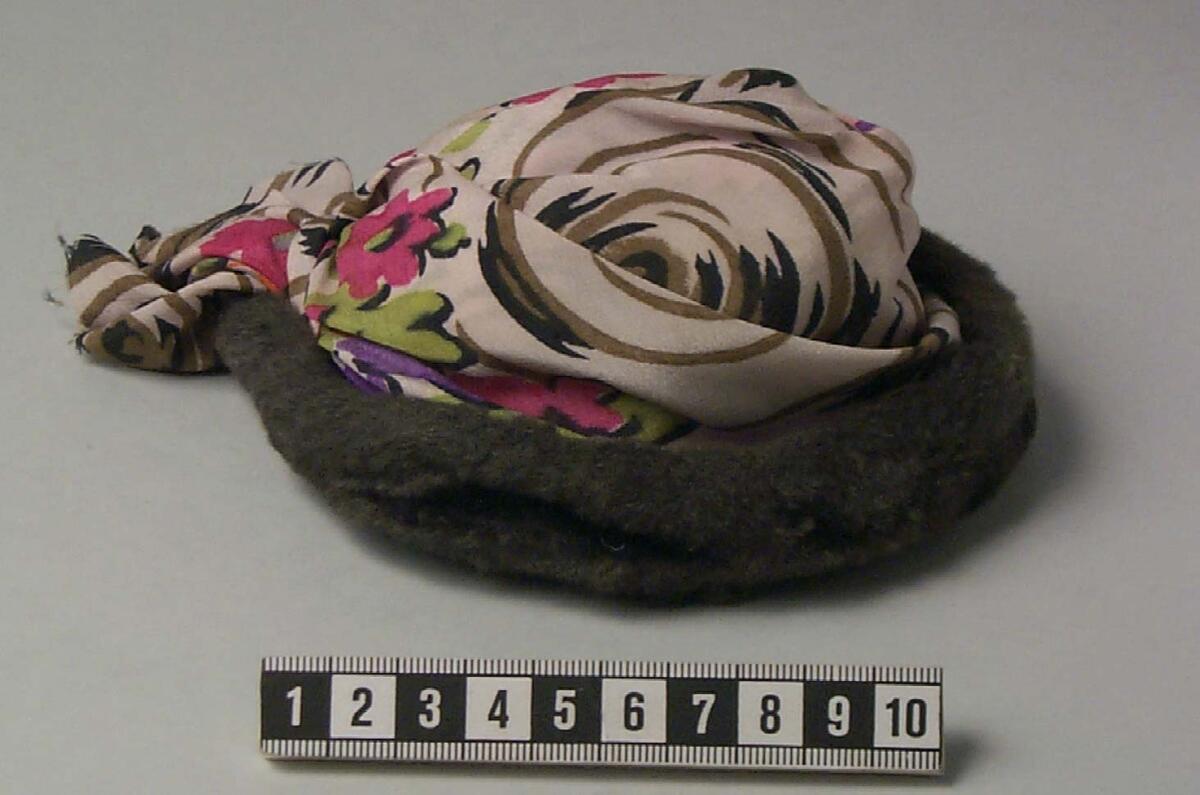 Rund hatt till docka, gråbrun skinnkant, kulle av blommigt siden i rosa grönt lila orange svart och beige mot ljus botten, en kort band av tyget hänger utanför kanten, fodrad med vitt tyg, inuti ett fastsytt band med text " i stuebecke", enligt originalkatalogen gåva av byråinspektör ingrid bohman, stockholm, syster till rektorn vid sävesskolan i visby, lennart bohman.
av Blommigt siden, gråbrun skinnkant. Inuti " i stuebecke".