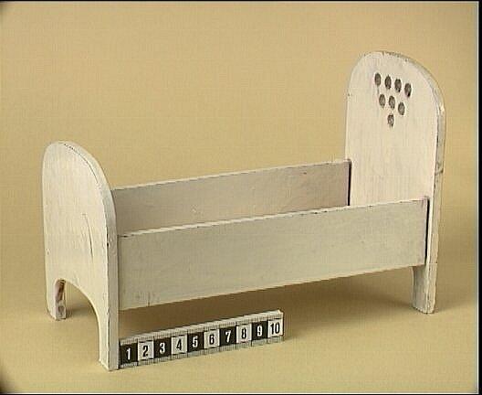 Vitmålad säng av trä, gavlarna har rundade överdelar, huvudgaveln är högre och har genomborrade hål som dekor, lös träskiva som botten, bäddad med sängkläder se inventnr c11172:137.