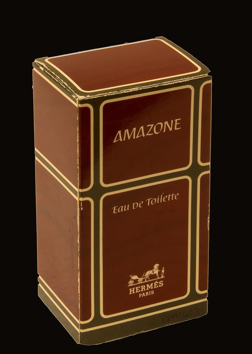 Parfymflaska, Amazone av Hermès, först lanserad 1974. Förvaras i originalförpackning. Flaska av glas med texten "AMAZONE Eau De Toilette HERMÈS PARIS" framtill. Lock av brunröd plast med marmorering. I flaskans botten texten "BOTTLE MADE IN FRANCE" samt ett klistermärke med texten "FACTICE". Flaskan innehåller vätska, men är en displayversion för butik och innehåller inte riktig parfym. Förpackning av papper med geometriskt mönster i rött, grönt och beige, med texten "AMAZONE Eau De Toilette HERMÈS PARIS" framtill. Del av parfymsamling från företaget Stengårds Färg som förutom färgmaterial även sålde parfym och skönhetsprodukter.
