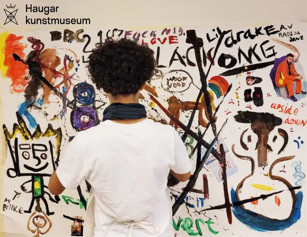 En person står med ryggen til. H*n har hvit skjorte og sort, krusete, hår. Personen ser på et abstrakt maleri