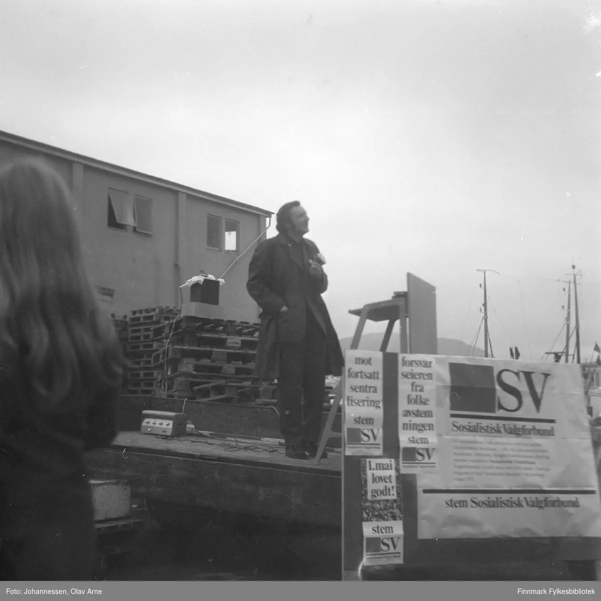 En mann står på scene og holder politisk tale på torget ved Havly og Langsethkiosken i Båtfjord, Finnmark

Foto trolig tatt på tidlig 1970-tallet

Foran han står skilt med teksten: mot fortsatt sentralisering stem SV, Forsvar seieren fra folkeavstemningen SV, 1. mai lovet godt! stem SV, SV sosialistisk Valgforbund, stem Sosialistisk Valgforbund 