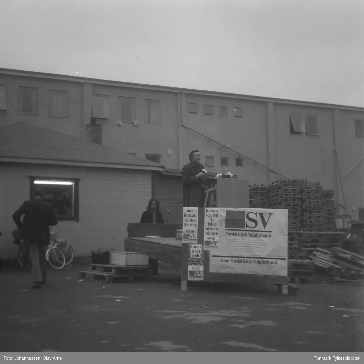 En mann står på scene og holder politisk tale på torget ved Havly og Langsethkiosken i Båtfjord, Finnmark

Foto trolig tatt på tidlig 1970-tallet

Foran han står skilt med teksten: mot fortsatt sentralisering stem SV, Forsvar seieren fra folkeavstemningen SV, 1. mai lovet godt! stem SV, SV sosialistisk Valgforbund, stem Sosialistisk Valgforbund 