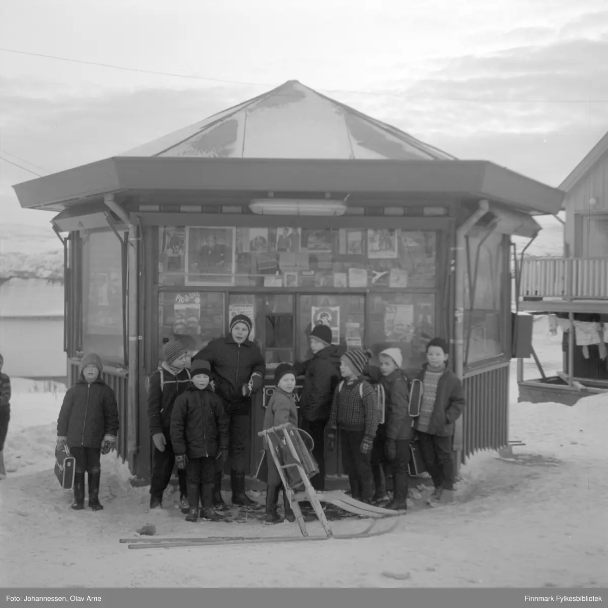 En gjeng gutter står foran Ragnvaldkiosken i Skolegata. Ragnvald Johansen i disken. 

Fra høyre Tommy Aasjord, Torfinn Rasmussen, Frank Sandberg (alle født i 1956), Arild Pedersen (ved luken), Kjell Otto Nilsen, tre siste er ukjente. 

Odd Jarle Jensen til venstre. Skolegata 25 til høyre.

De er kledd i vinterklær, jakker og luer. 

Til høyre kan man se en parkert spark og i bakgrunnen ser man bolighus 