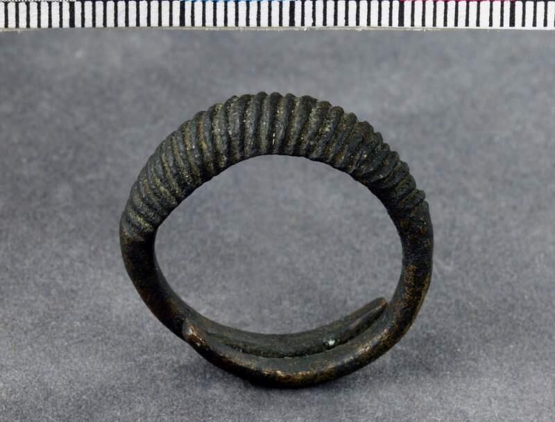 Ring av brons, öppen modell, de avsmalnande ändarna lagda omlott, "framsidan" tjockare och välvd samt ornerad med täta snedställda linjer
av brons, största tvärmått 29 mm.