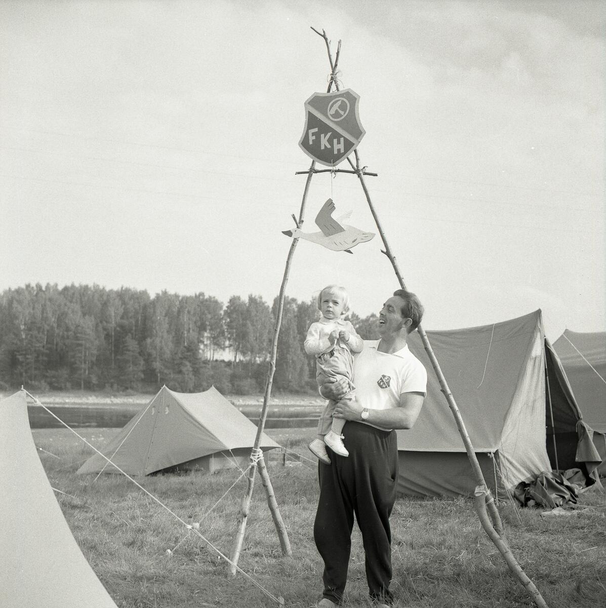 En man håller i ett barn vid entrén till ett tältläger under Svenska Frisksportförbundets riksläger i Bollnäs den 12-19  juni 1959.